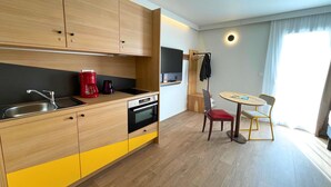 Une cuisine moderne avec des meubles en bois, un évier, une machine à café rouge et un four encastré. On y trouve également un coin repas avec une table et des chaises, ainsi qu’un téléviseur fixé au mur.
