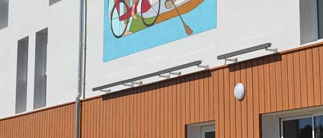 En byggnad med en stor väggmålning som föreställer människor på en cykel och i en båt, en träfasad och en klarblå himmel.