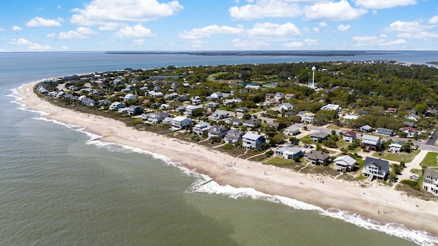 Vista aérea de una zona residencial costera con casas a lo largo de la playa, un faro y una vista despejada del océano.