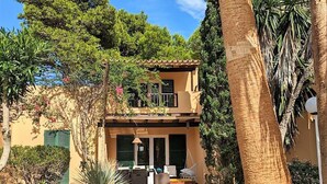 Une maison à deux étages dotée d’un balcon, entourée d’une végétation luxuriante et de palmiers.