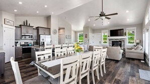 A spacious kitchen with modern appliances, a dining area with white chairs, and a living room with a fireplace. - Bear Lake Getaway |Sleeps 19, Hot Tub, Views, Ideal Beach, Ping-Pong! (Garden City)