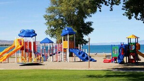 A playground with various play structures including slides and climbing areas, set against a backdrop of a clear sky and a body of water. - Bear Lake Getaway |Sleeps 19, Hot Tub, Views, Ideal Beach, Ping-Pong! (Garden City)