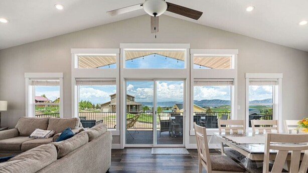 A spacious living room with a large window, a ceiling fan, and a dining area with a view of the outdoors. - Bear Lake Getaway |Sleeps 19, Hot Tub, Views, Ideal Beach, Ping-Pong! (Garden City)
