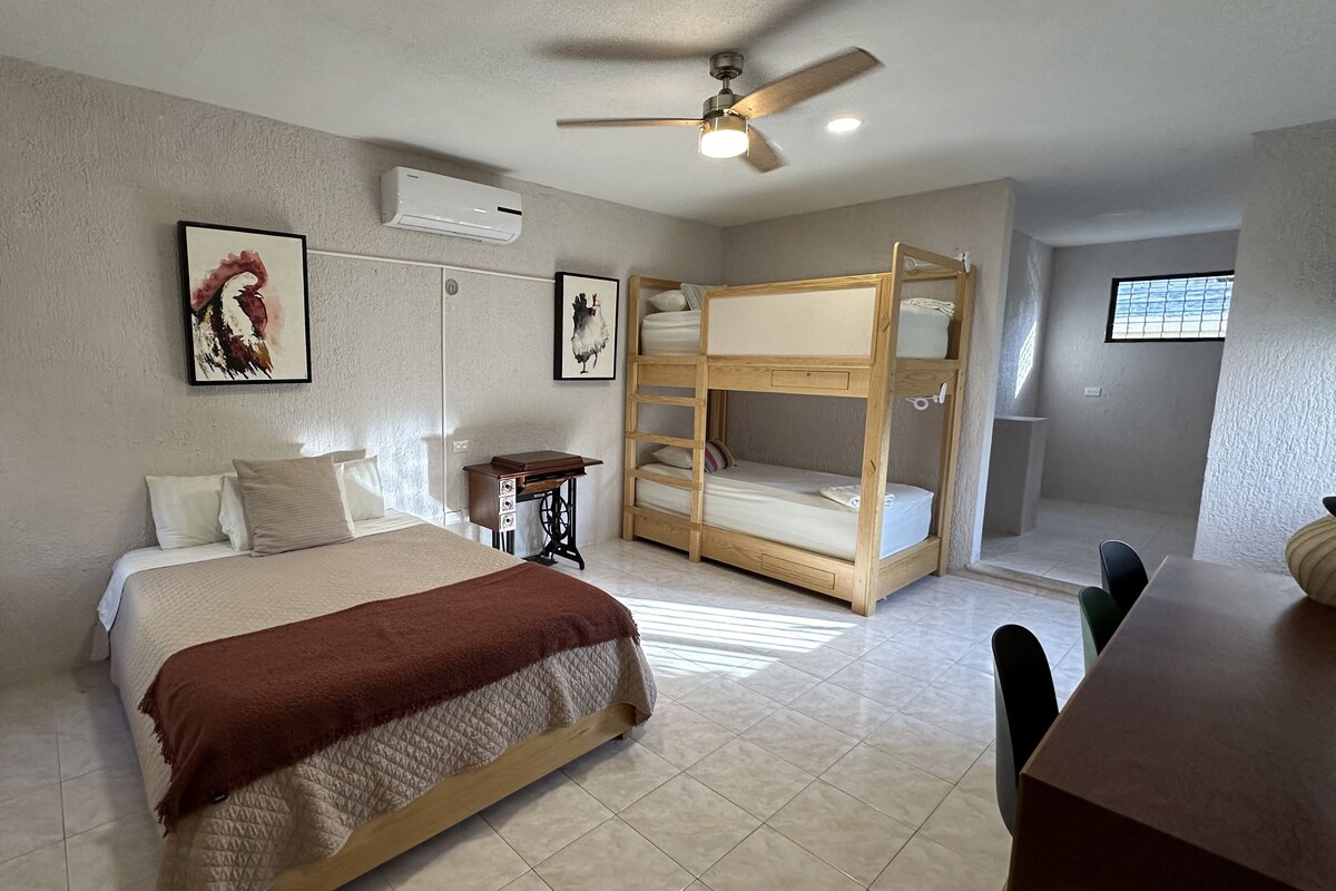 Une chambre à coucher comprenant un lit, un lit superposé en bois, un bureau, une chaise, un ventilateur de plafond et des œuvres d’art accrochées aux murs.