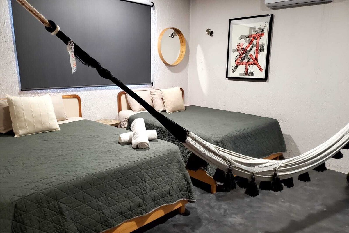 Une chambre avec deux lits, un hamac, un miroir rond et une œuvre d’art encadrée au mur.