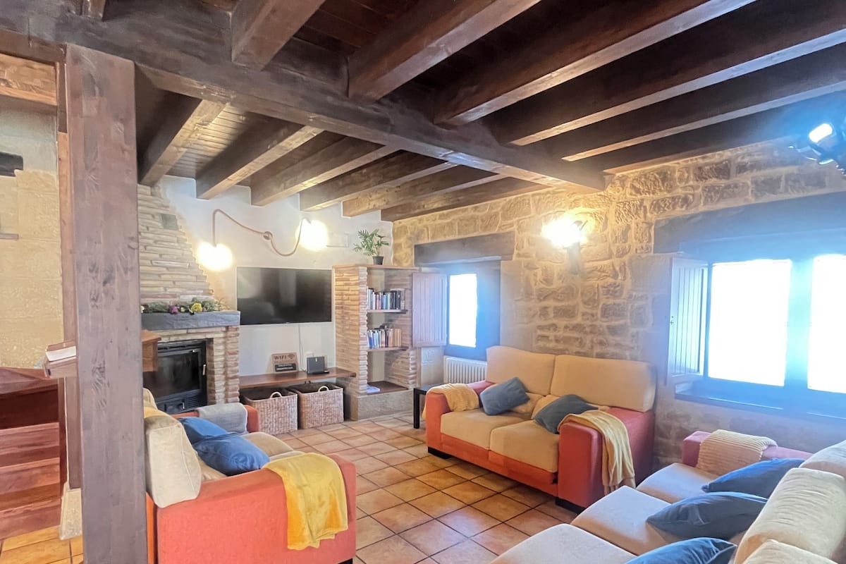 A cozy living room with a fireplace, stone walls, and wooden beams. There are sofas and a coffee table with cushions.
