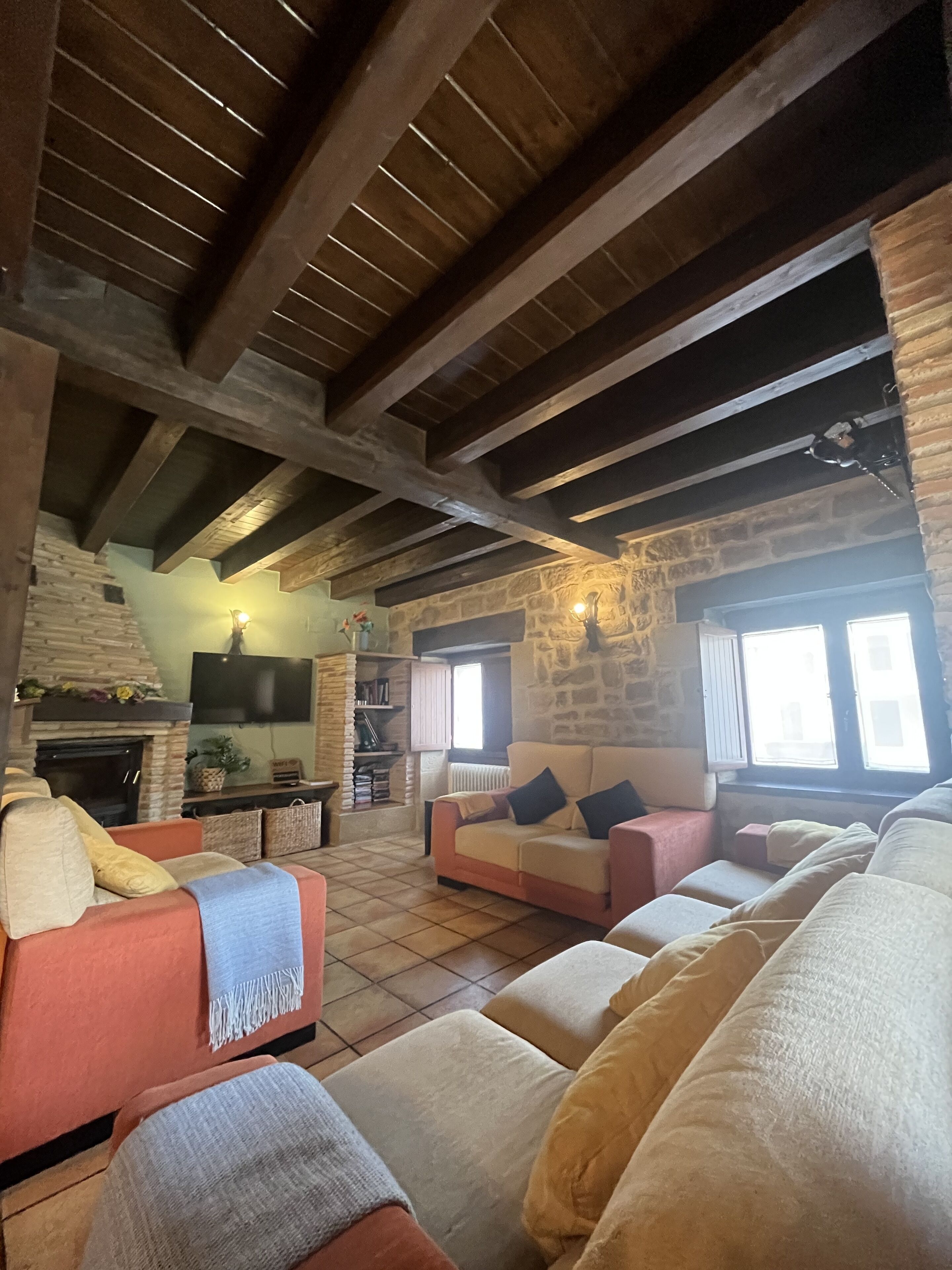 A cozy living room with a fireplace, stone walls, and wooden beams. There are sofas and a TV mounted on the wall.