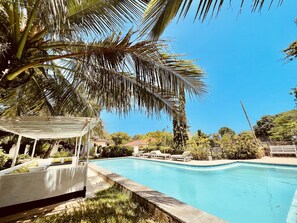 Une piscine dotée d’un espace de repos couvert, entourée de palmiers et surplombée d’un ciel bleu azur.