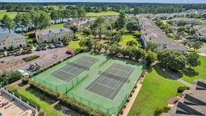 An aerial view of a residential area with two tennis courts, a parking lot, and houses with a golf course in the background. - Legends Golf Condo Steps From Ailsa Pub & Pool (Myrtle Beach)