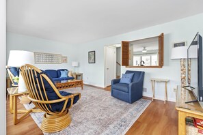 Ein Wohnzimmer mit einem blauen Sofa, einem Korbstuhl, einem kleinen Tisch und einer TV-Konsole.