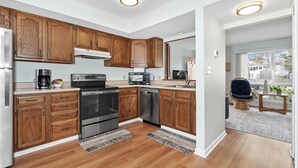 A modern kitchen with wooden cabinets, stainless steel appliances, and a view to the living area. - 215 Cedar Street Unit 4 Rose Garden - Lewes Beach (Lewes)