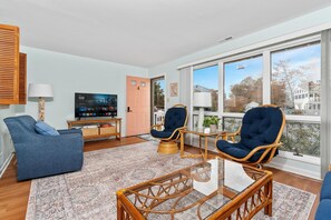 Ein geräumiges Wohnzimmer mit einem großen Fenster, einem blauen Sofa, zwei Korbsesseln, einem Glastisch und einem Flachbildfernseher.