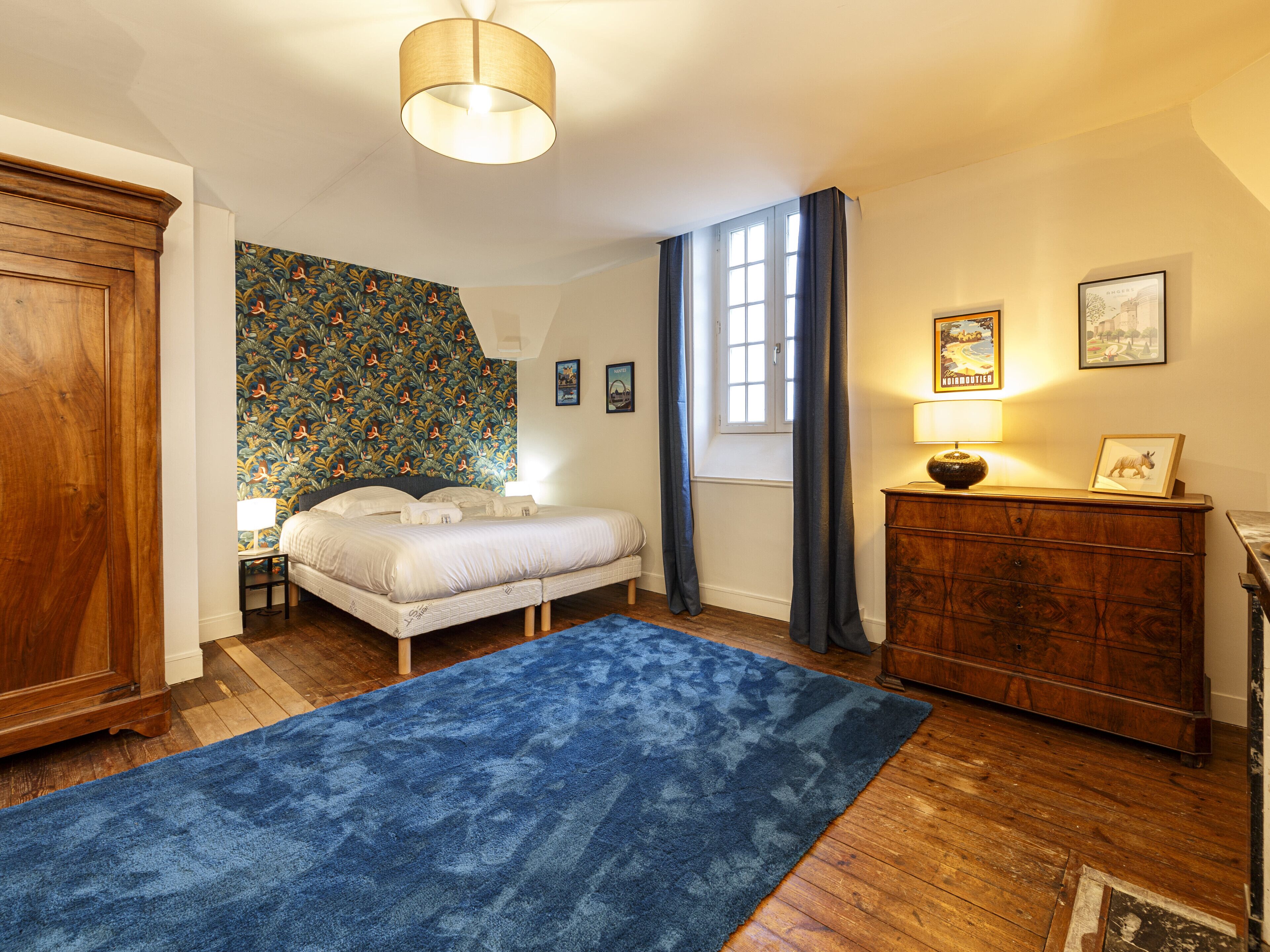 A bedroom with a bed, a dresser, a nightstand, a lamp, a window with curtains, and a wall with a floral wallpaper.