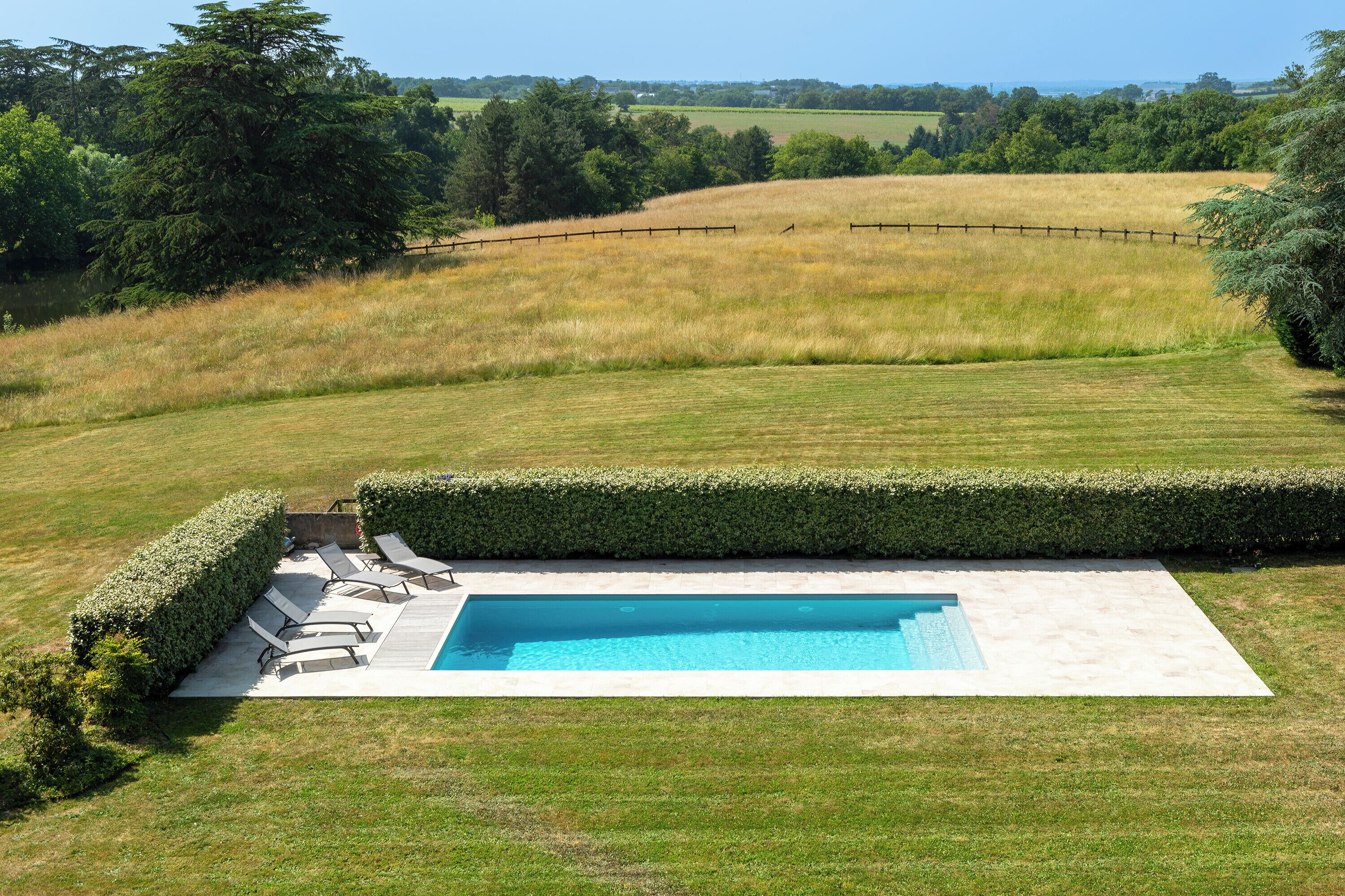 Ein rechteckiger Pool mit Holzterrasse und Liegestühlen, umgeben von einem gepflegten Rasen und einer Hecke.