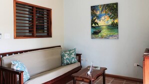 Ein Zimmer mit einer hölzernen Sitzbank, einem Sofa mit gemusterten Kissen, einem kleinen Tisch und einem Bild einer tropischen Szene.