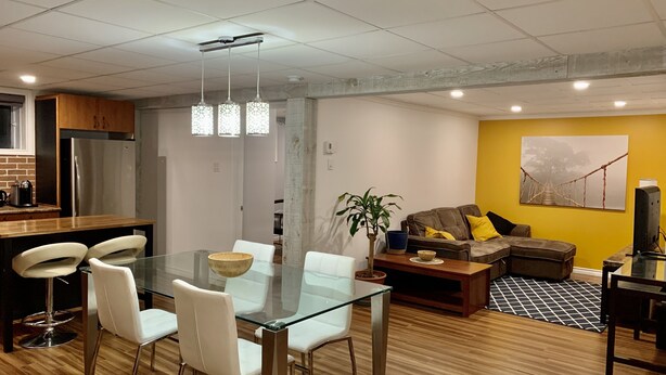 A modern living space with a dining area featuring a glass table and white chairs, a kitchen with stainless steel appliances, and a cozy seating area with a sofa and cushions. - The Bachelor (Mirabel)