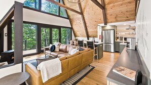 A spacious loft with a high ceiling, a cozy seating area with a sofa and chairs, a kitchen area, and large windows offering a view of the outdoors. - 1 mile to Bryce Resort l 4 BR l Hot Tub l Fire Pit l Game Room  (Basye)