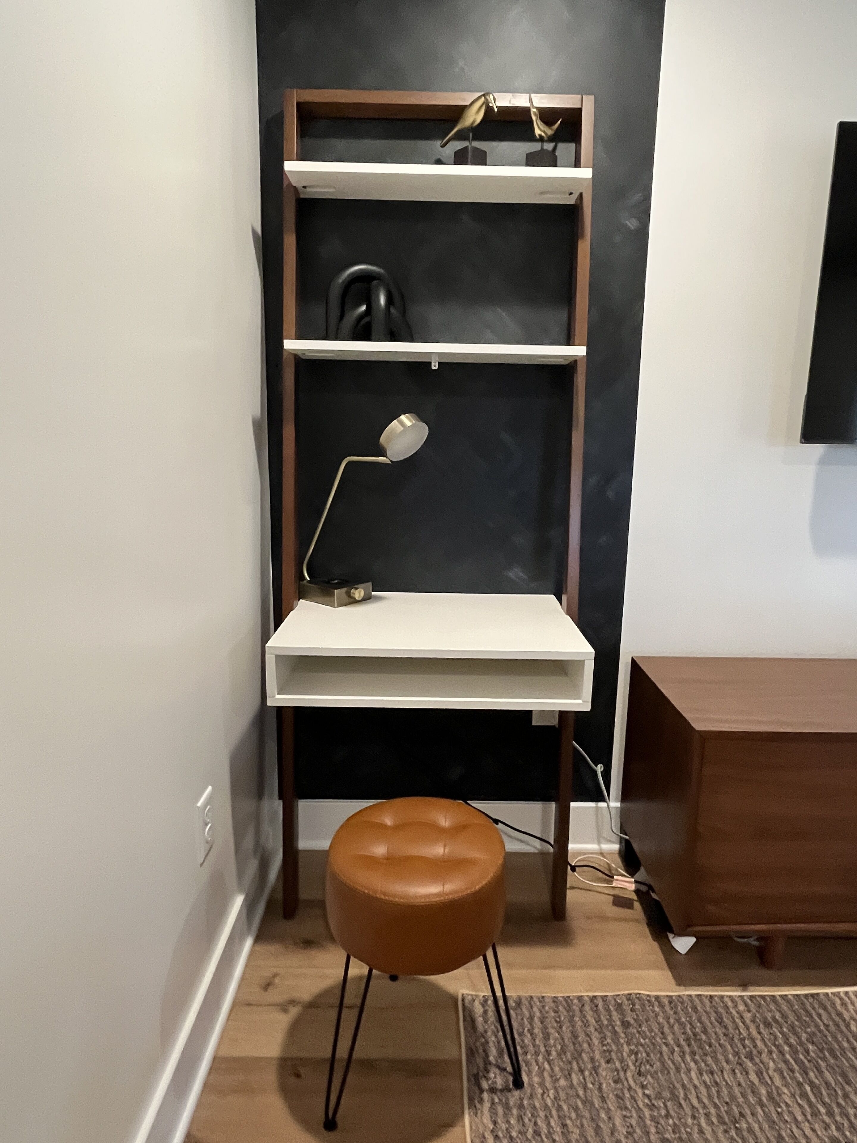 A compact workspace with a desk, shelves, a lamp, and a brown stool.