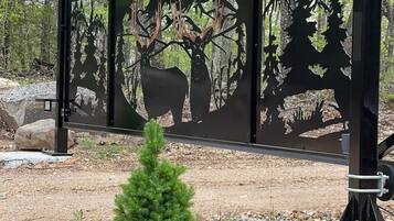 Una cerca metálica con un corte decorativo de paisaje forestal, rodeada de rocas y un pequeño árbol de hoja perenne.