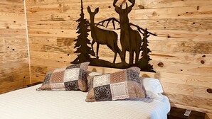 A wooden cabin room with a bed, patterned pillows, a wall-mounted air conditioner, and a decorative deer and tree cutout. - Country Cabin with Jacuzzi Tub and Meramec River Access and Pets welcome! (Bourbon)