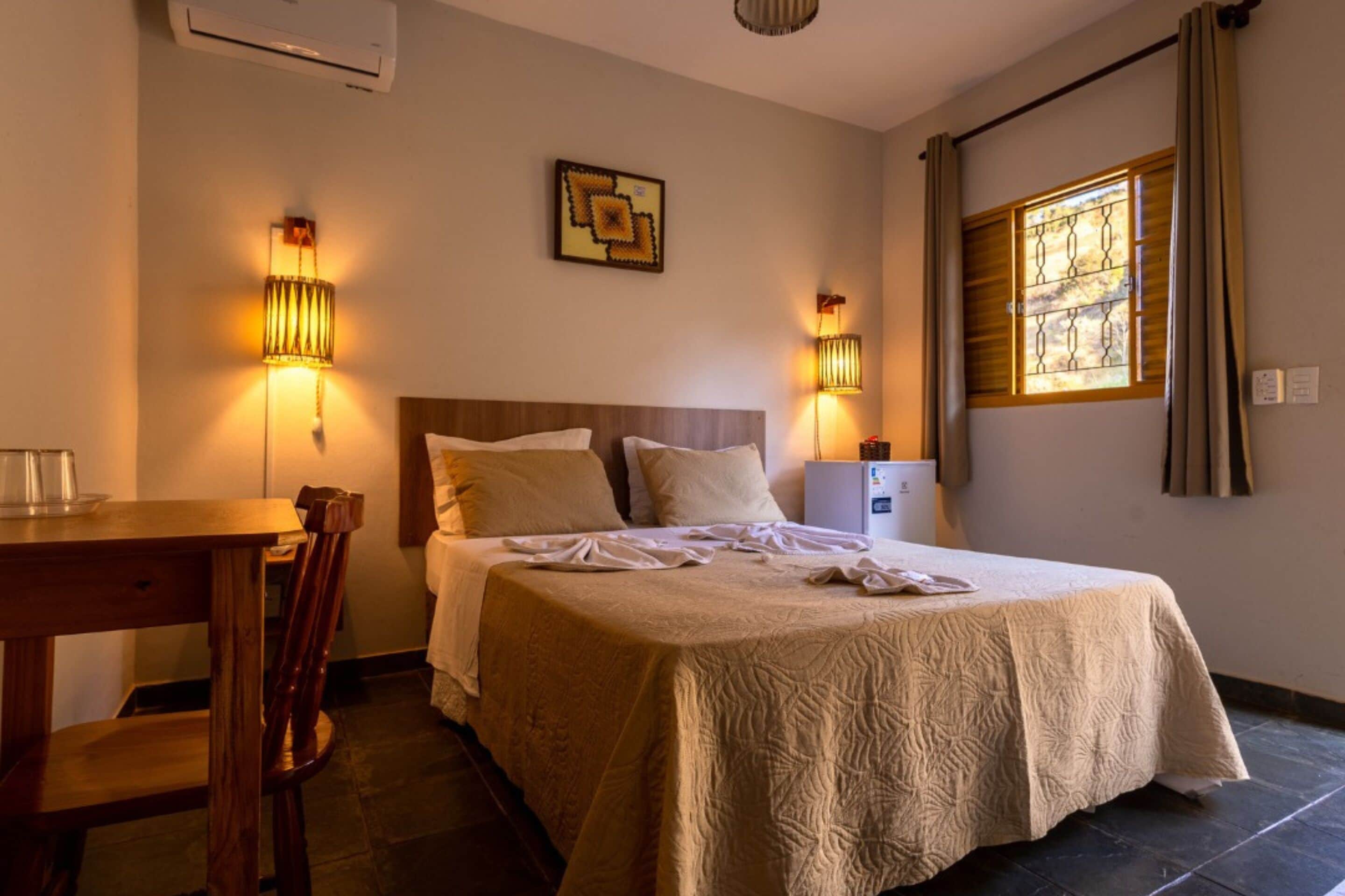 Quarto de hotel com cama, luminárias de cabeceira, escrivaninha e janela com cortinas.