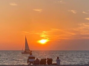 A sunset over the ocean with a sailboat and beach chairs.