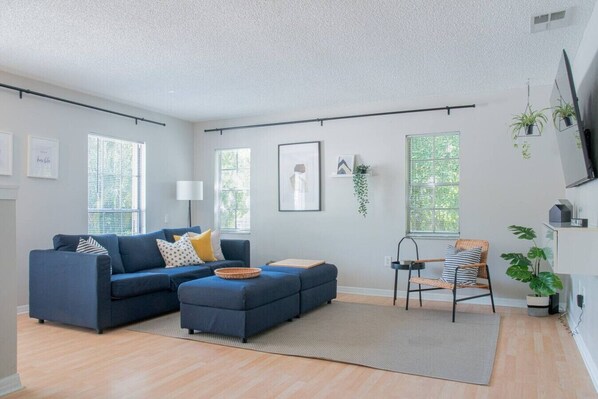 A modern living room with a blue sofa, a small ottoman, a wicker chair, and a flat-screen TV mounted on the wall.