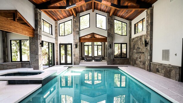 Una amplia piscina interior con paredes de piedra y vigas de madera, rodeada de grandes ventanales.