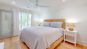 Un dormitorio con una cama grande, un ventilador de techo, una mesita de noche con lámpara y una ventana con cortinas.