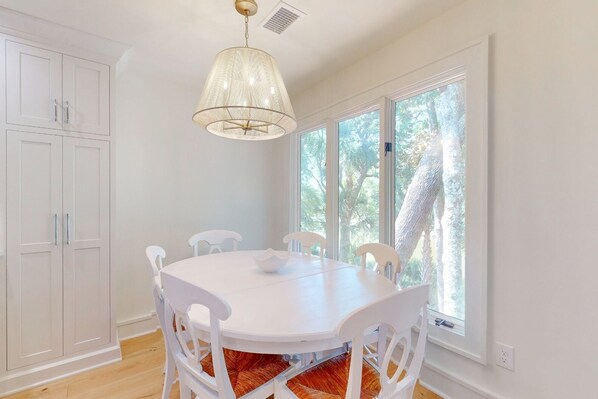 A dining area with a white table and chairs, a hanging light fixture, and large windows with a view of trees.