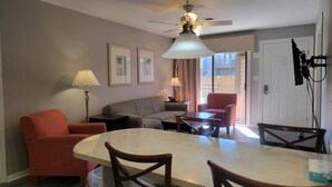 A dining area with a table, chairs, and a ceiling fan. There are two framed pictures on the wall, a lamp, and a door with a lock.