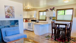 Cozinha moderna com armários brancos, micro-ondas e fogão. Possui uma área de jantar com mesa e cadeiras de madeira, e uma sala de estar com sofá azul e mesa de centro de vidro.