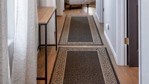 Een gang met behang, een houten bank en een vloerkleed dat leidt naar een slaapkamer met een bed, lamp en raam.