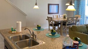 A modern kitchen with granite countertops, stainless steel sink, and pendant lighting. There is a dining area with a table and chairs, and a view of the outside through a window. - Compass Bay by CozyKey (Kissimmee)