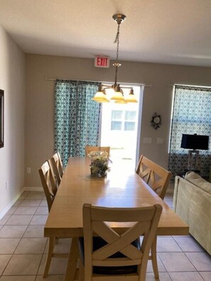 A dining area with a wooden table, chairs, and a chandelier. There is an exit sign above the door. - Compass Bay by CozyKey (Kissimmee)