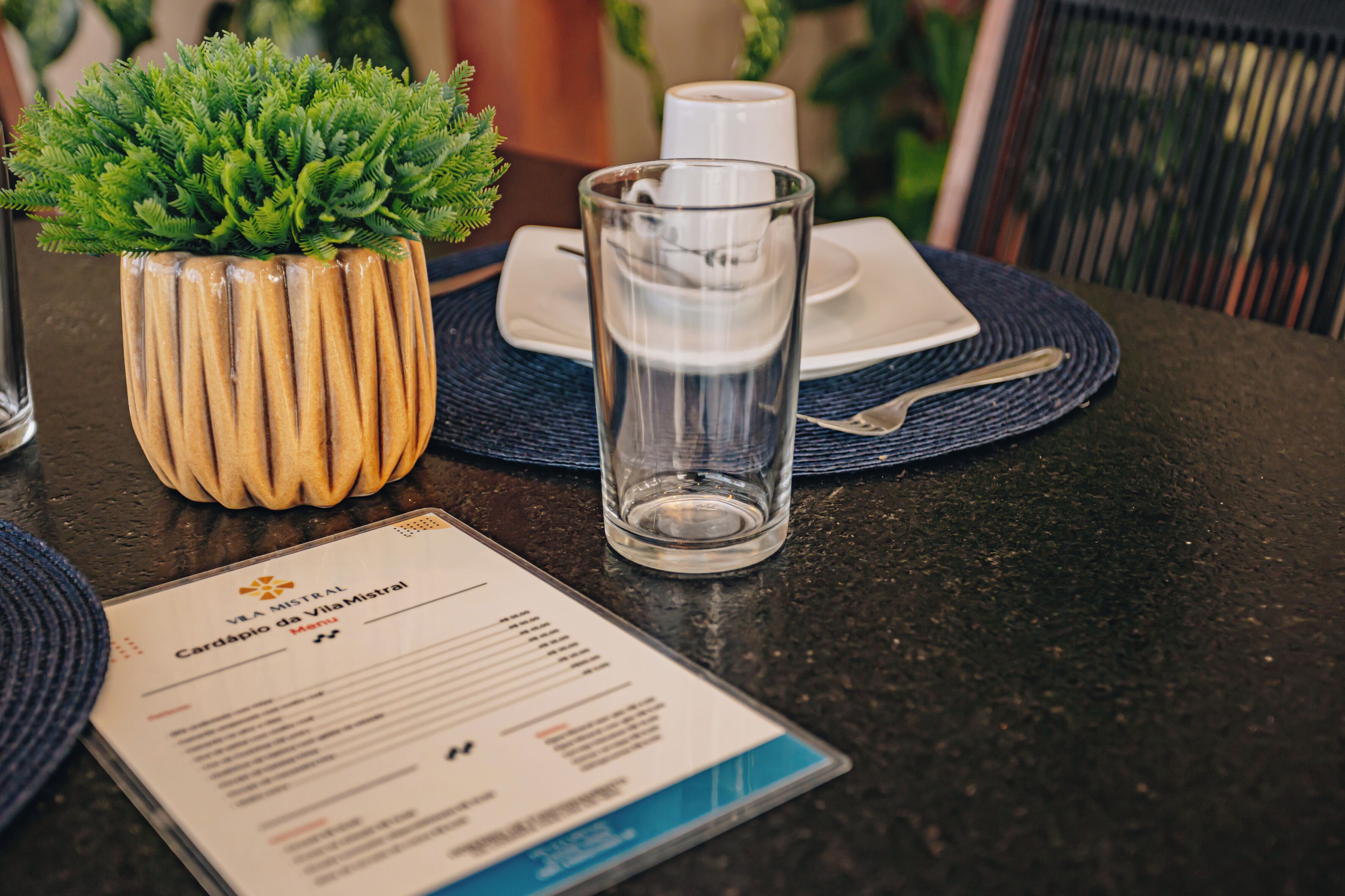 A menu on a table with a glass of water, a potted plant, and a cup.