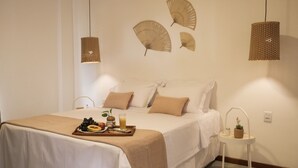 Una cama bien tendida con una bandeja de fruta y jugo, una mesita con una planta y tres ventiladores en la pared.