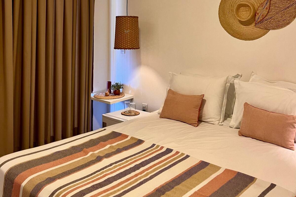 A hotel room with a bed, curtains, a wall-mounted air conditioner, a woven lamp, and a decorative wall piece.