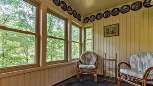 Une pièce dotée de grandes fenêtres, de chaises en osier, d’un tableau encadré et d’une étagère fixée au mur.