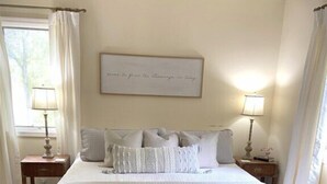 Ein ordentlich bezogenes Bett mit einer gesteppten Tagesdecke und mehreren Kissen, ein Nachttisch mit Lampe sowie ein gerahmter Spruch an der Wand.