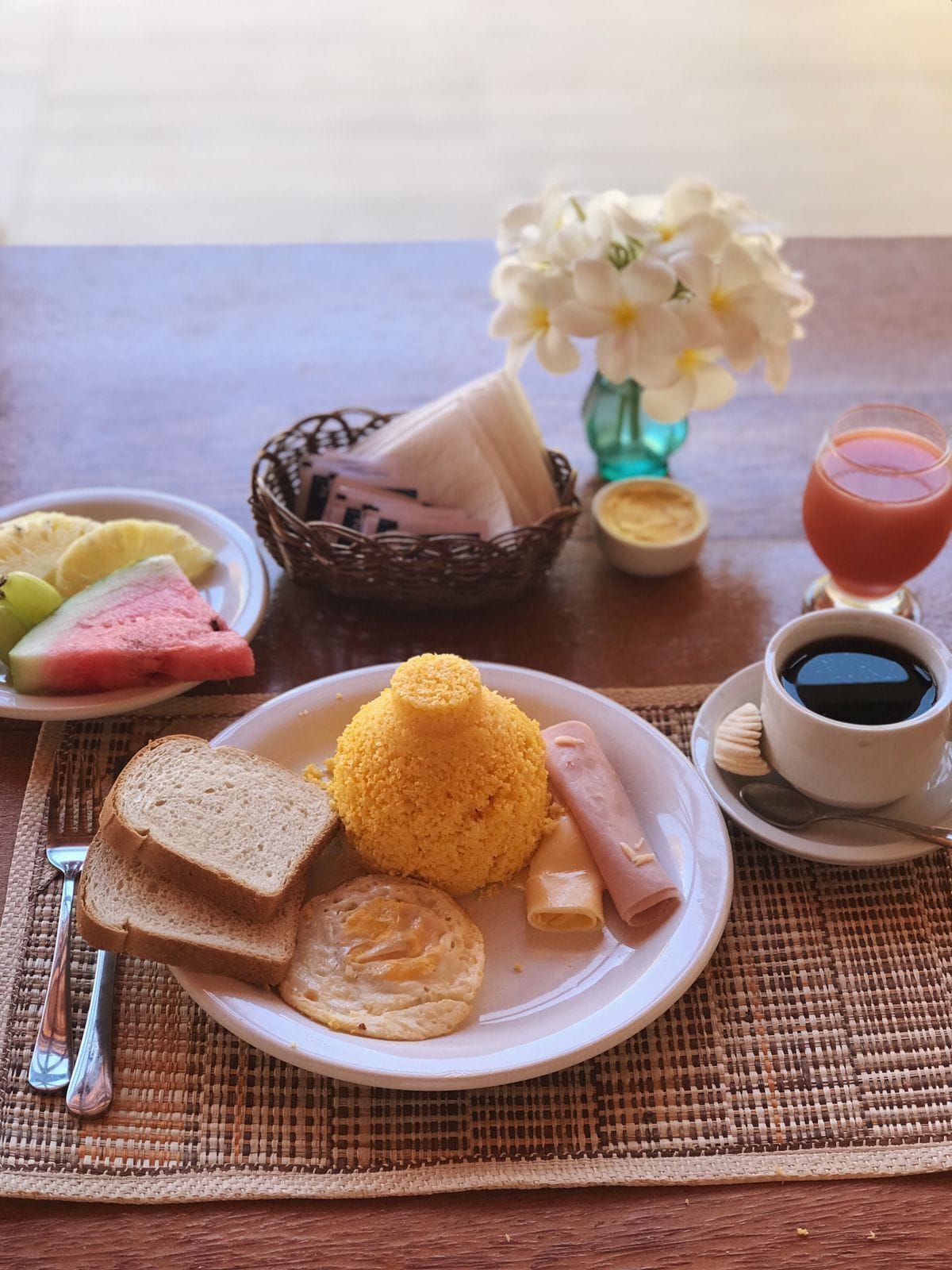 Une table à manger avec une assiette de nourriture, un panier, un vase de fleurs et une tasse de café.