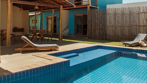 A pool area with a wooden deck, lounge chairs, and a two-story building with a blue exterior. - Pousada Villa Kai  (São Miguel dos Milagres)