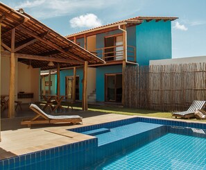 Ein Poolbereich mit Holzterrasse, Liegestühlen und einem zweistöckigen Gebäude mit blauer Fassade.