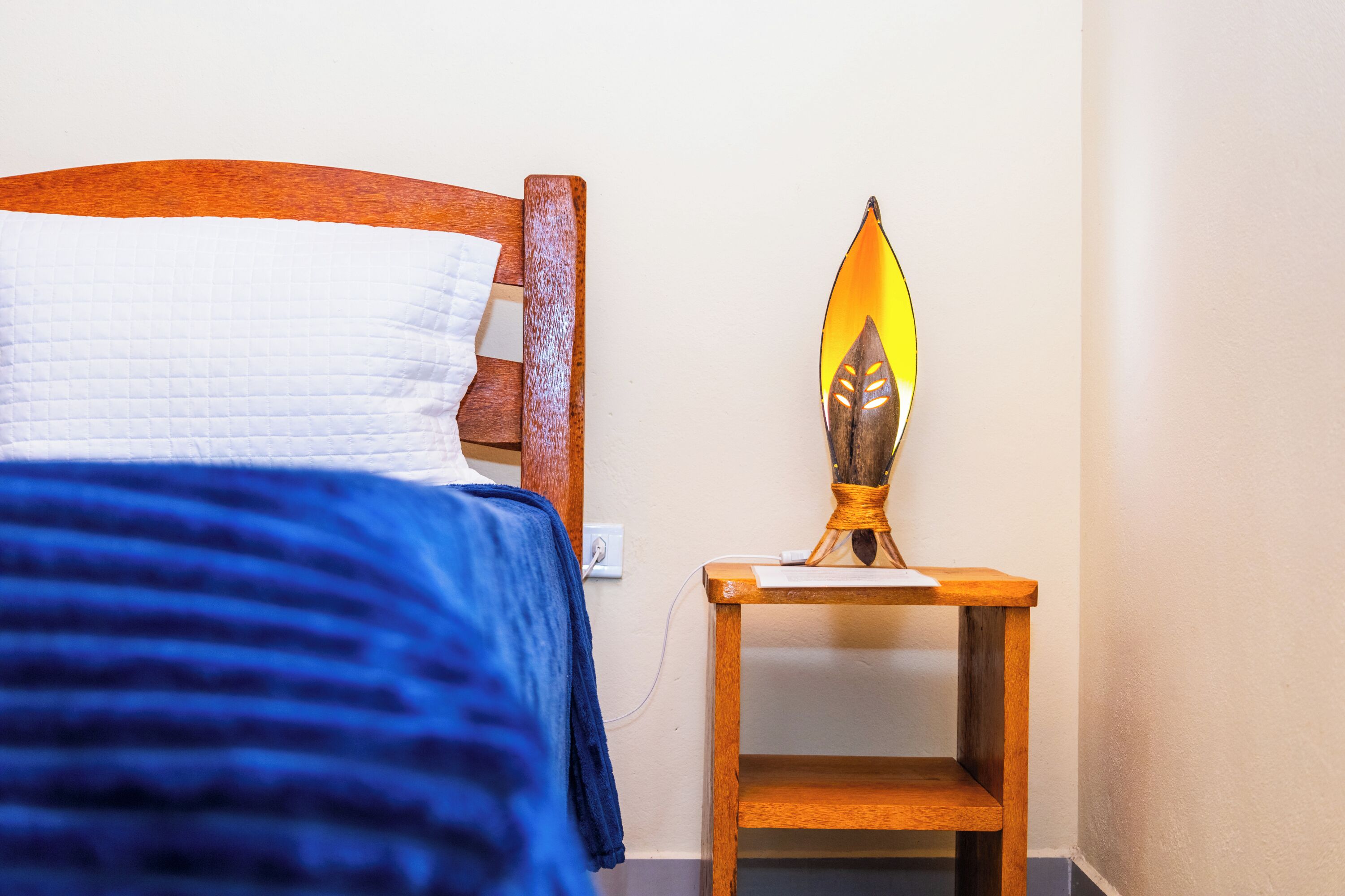 A bed with a blue blanket and a wooden headboard, a bedside table with a lamp, and a power outlet on the wall.