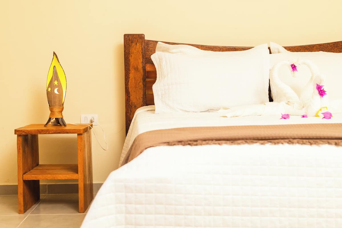 Un lit bien fait, recouvert d’une couette blanche et d ’une couvre-lit marron, une table de chevet en bois avec une lampe, et un mur au décor sobre.