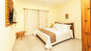 A hotel room with a bed, a small wooden stool, a nightstand with a lamp, and a wall-mounted TV. - Pousada Villa Kai  (São Miguel dos Milagres)