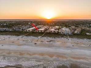An aerial view of a coastal area with buildings, a beach, and a sunset.