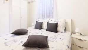 Een slaapkamer met een bed, kussens, een nachtkastje, een spiegel, een kledingkast en een raam met gordijnen.