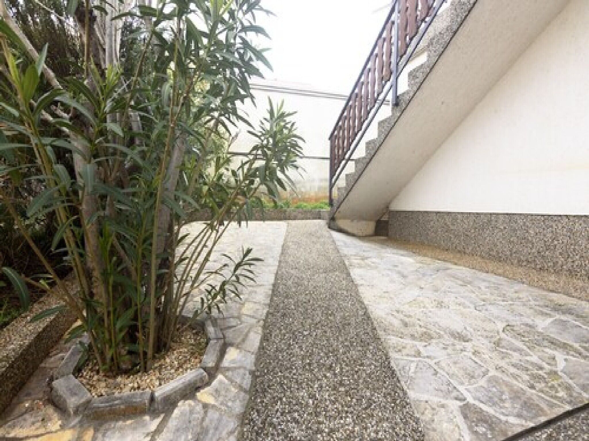 Ein gepflasterter Weg mit Mittelstreifen, gesäumt von Pflanzen und einem Gebäude mit weißer Fassade und Steinsockel.