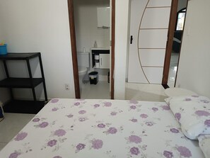 Cozy retreat with a floral-patterned bedspread and a wooden-framed door leading to a bathroom with a toilet, sink, and trash bin.